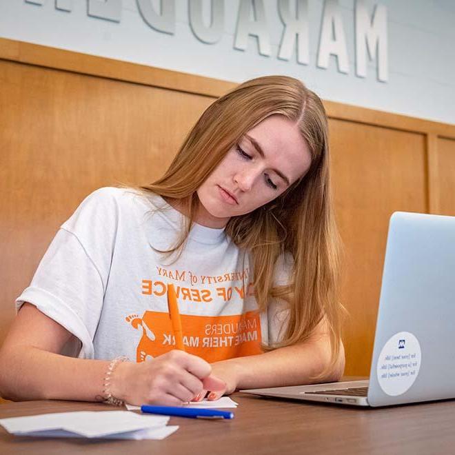 Female student writing notes with laptop on table in front of her.